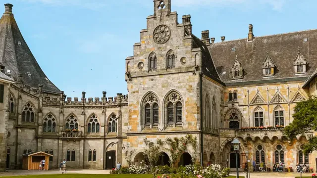 Historic stone building with clock tower, surrounded by gardens, under a blue sky.