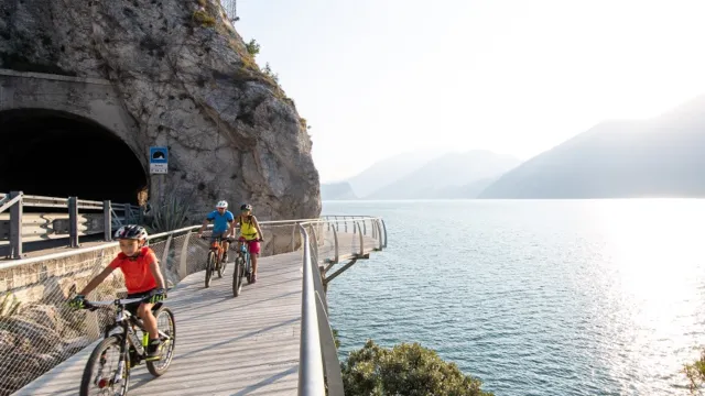 Cyclists ride on a lakefront path near a mountain tunnel at sunset.