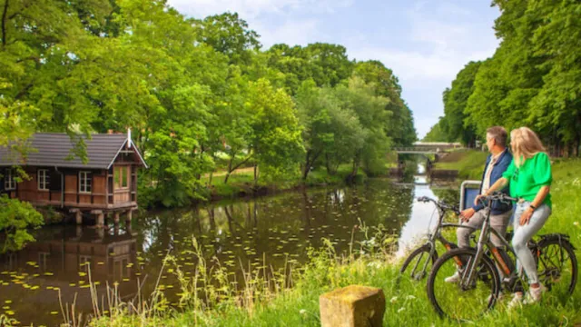 Two people with bikes admire a riverside park with lush greenery and a wooden cabin.