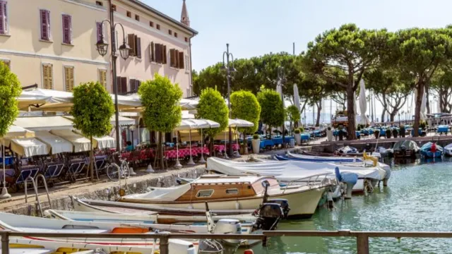 Boats docked at a quaint waterfront lined with cafes and trees under a blue sky.