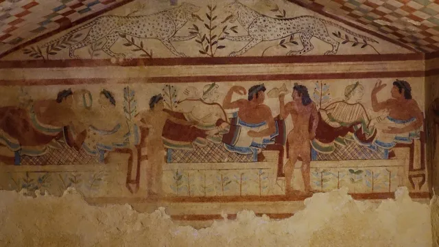 Ancient fresco depicting figures reclining at a banquet, with geometric ceiling patterns.