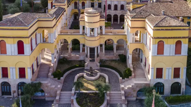 Elegant villa with yellow walls and arches, surrounded by greenery and flags.