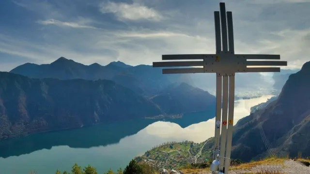 Metal cross on a mountain peak overlooking a tranquil lake and distant hills.