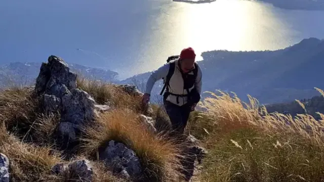 Hiker on rocky trail with sea view, sun reflecting on water.