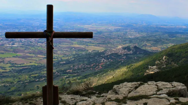 Cross on a rocky hilltop overlooking a vast valley under a cloudy sky.