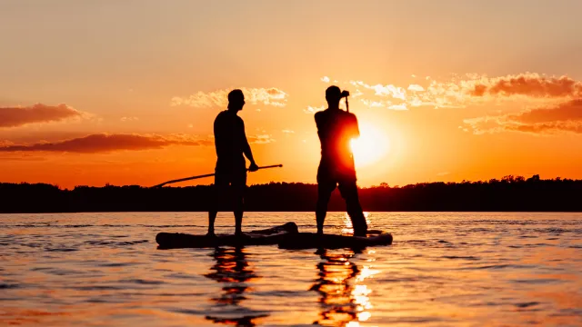Two people paddleboarding on a lake at sunset.