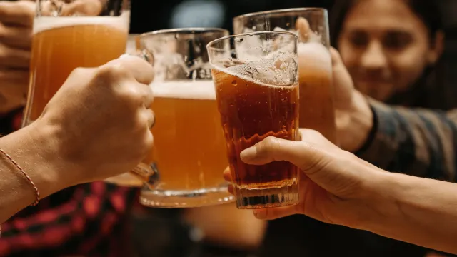 Four people clinking beer mugs over a wooden table in a dimly lit bar.