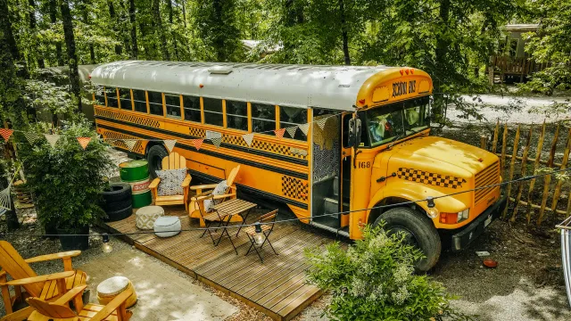 Yellow school bus converted into a cozy outdoor living space in a wooded area.