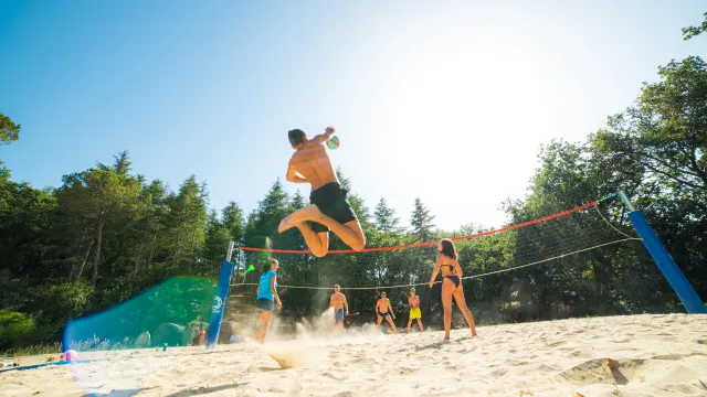 Beach volleyball game under clear skies, player mid-jump, surrounded by trees.