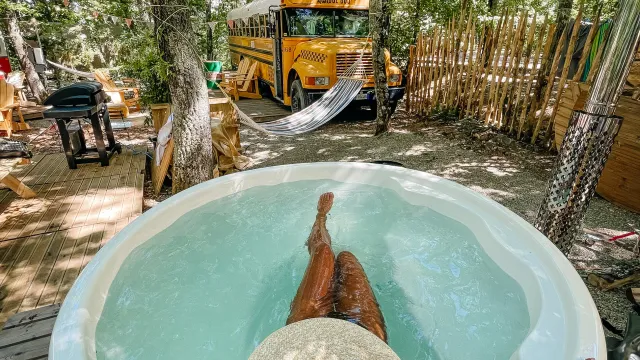 Person relaxing in a hot tub, wearing a straw hat, with trees and yellow bus in the background.