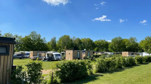 Grass field with camper vans, wooden cabins, and trees under a blue sky.