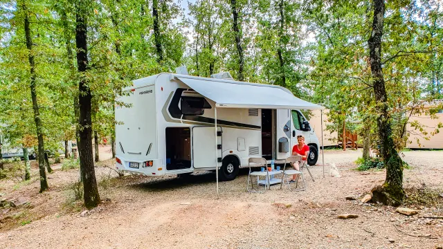 Camper van parked in a wooded campsite with chairs and table outside.