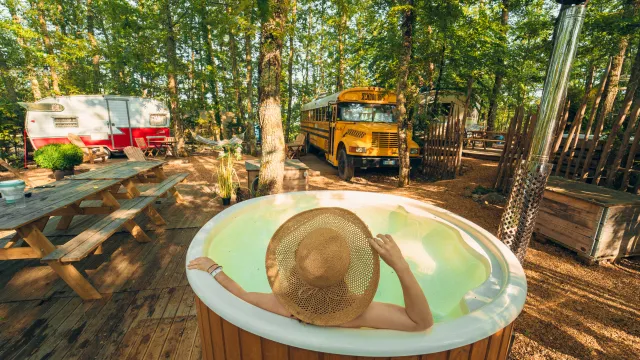 Person in a hot tub wearing a sun hat, surrounded by trees, camper, and a school bus.