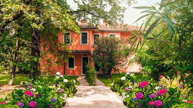 Pathway leads to a quaint red house surrounded by lush trees and colorful flowers.