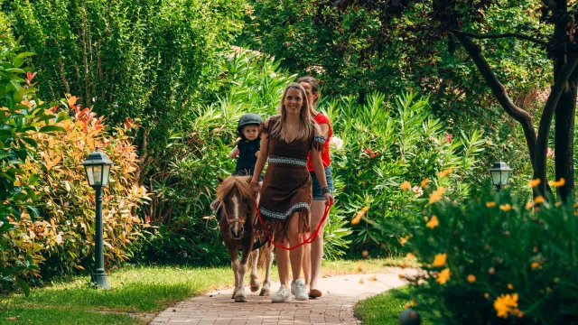 Child riding a pony with two adults guiding in a lush garden path.