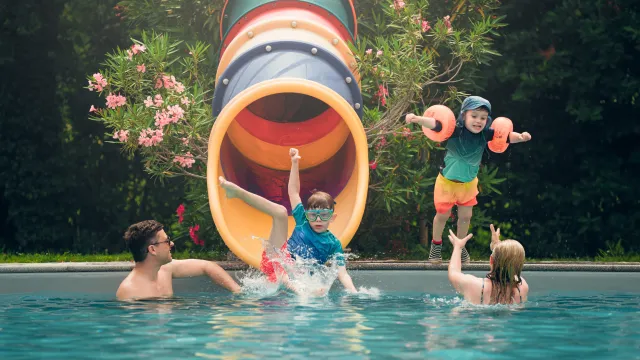 Children sliding into pool, adults catching them, colorful slide in background.