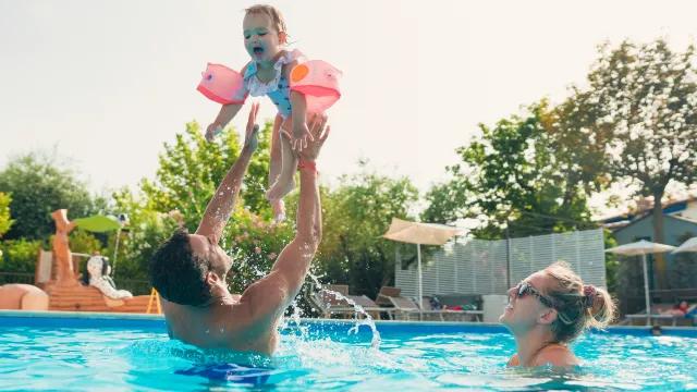 Father lifts child in pool with mother nearby, sunny day.