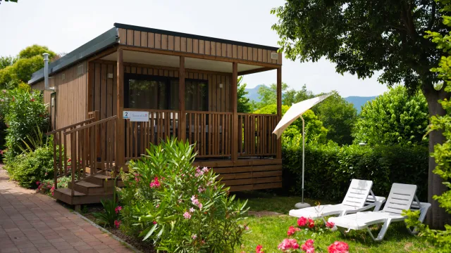 Cozy wooden cabin with a porch, surrounded by greenery and flowers; two lounge chairs nearby.