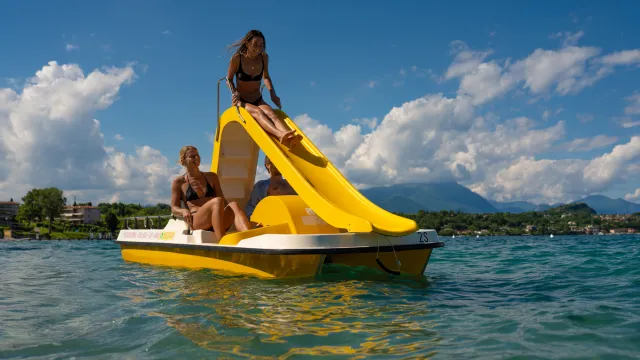 Two people on a pedal boat with a yellow slide in a sunny lake setting.