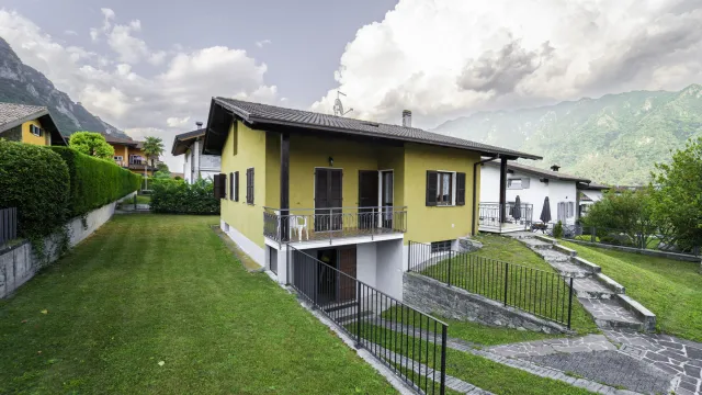 Yellow house with a sloped lawn, mountain backdrop, and a cloudy sky.