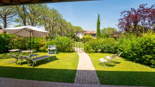 Sunny garden with lounge chairs, umbrella, and a path leading to trees and shrubs.