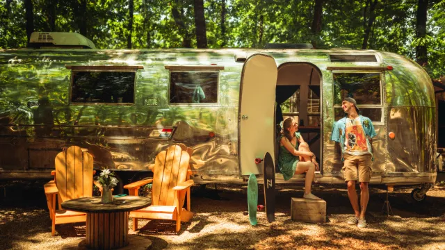 Retro camper surrounded by trees, two people relaxing outside.