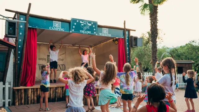 Children dancing in front of a small outdoor stage during the day.