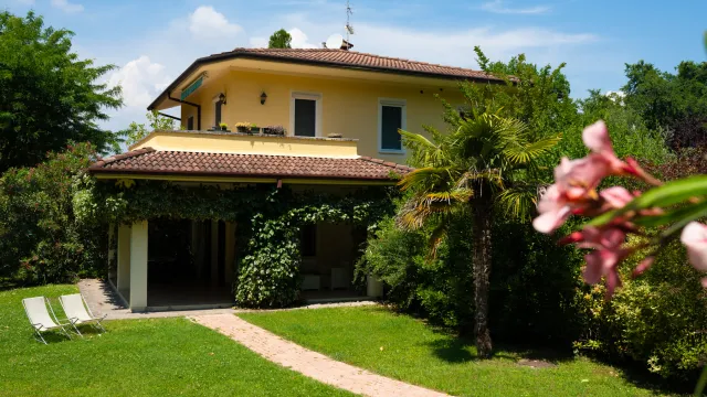 Two-story yellow house with garden, trees, and a small path under a blue sky