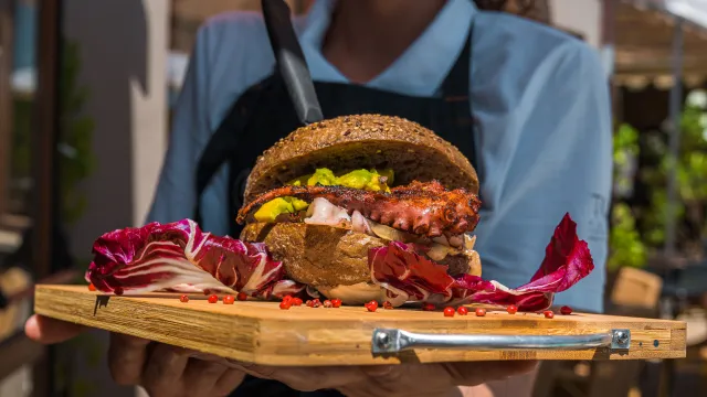 Smiling server holds a gourmet burger on a wooden platter outdoors.