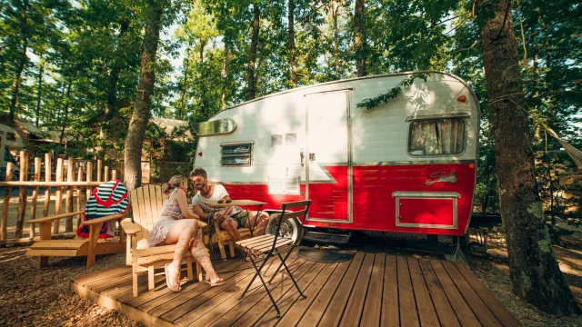 Camper in a wooded area with people sitting on a deck.