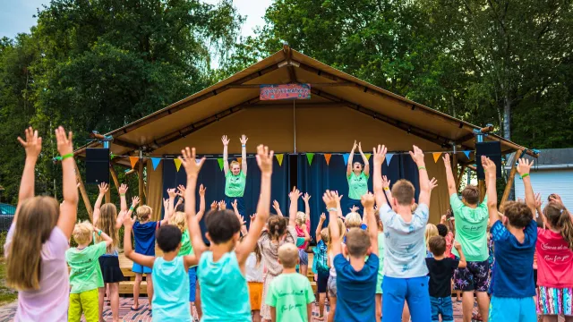 Children raise arms in unison at outdoor event in front of a stage.