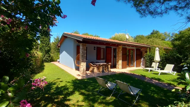 Small house with terrace in a sunny garden, surrounded by trees and flowers.