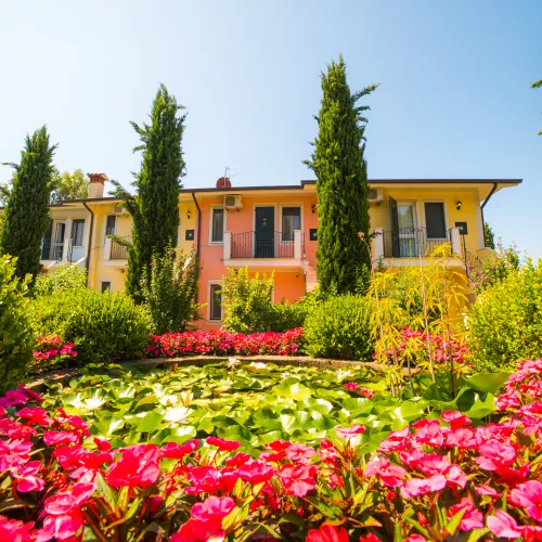 Colorful flowers and tall trees in front of a pastel villa under a clear blue sky.