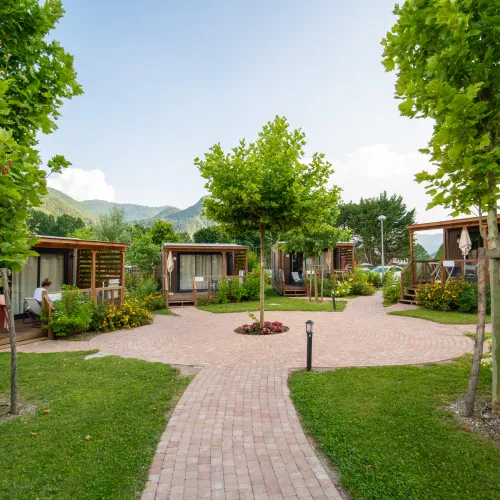 Pathway leading through a green park with cabins and trees against a mountain backdrop.