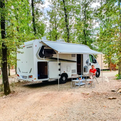 Camper van parked in a wooded campsite with chairs and table outside.