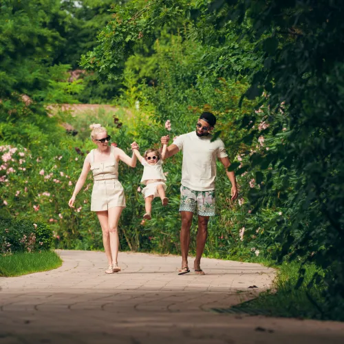Family walking on a path in a lush, green park, lifting a child between them.