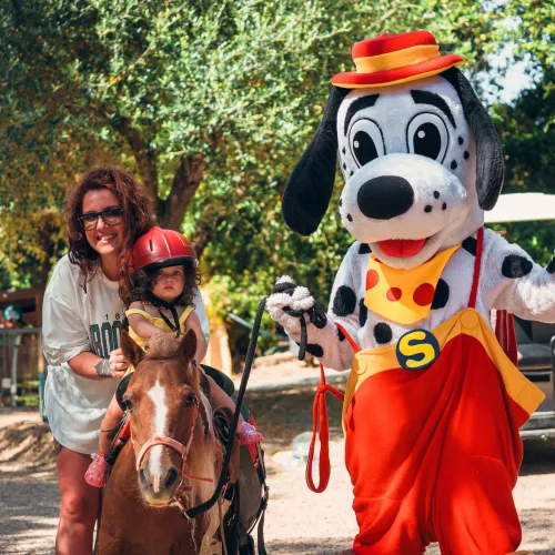 Child rides a pony with a woman; Spotty waves nearby.