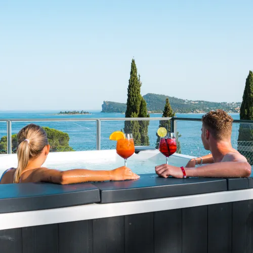 Couple relaxing in a hot tub with cocktails, overlooking a scenic lake and trees.