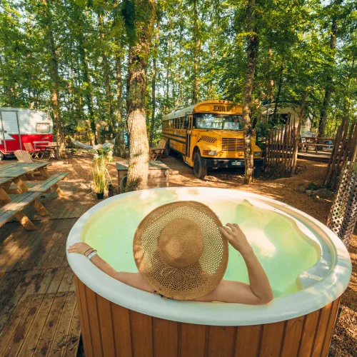 Person in a hot tub, wearing a straw hat, in a forest campsite with a bus and caravan.