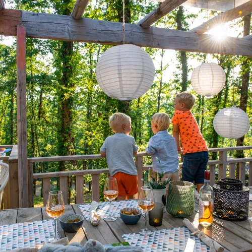 Children on a sunlit deck, surrounded by trees and lanterns, enjoying a late afternoon.