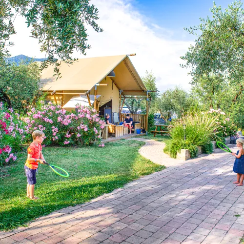 Children playing with rackets near a cottage, surrounded by lush greenery and flowers.
