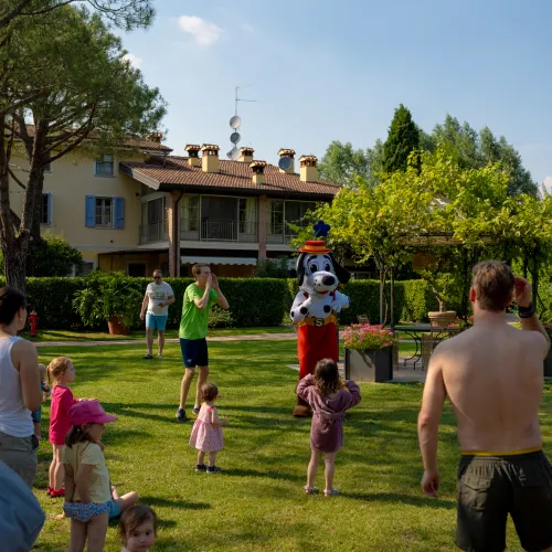 Adults and children gather on a grassy lawn, with trees and a house in the background.