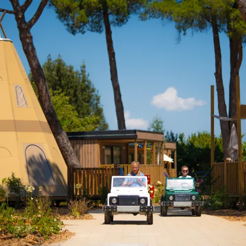 Two mini cars on a path near a tipi lodge, under tall trees.