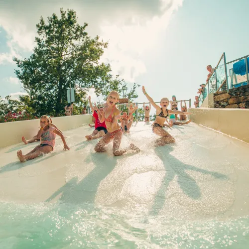 Children sliding down a water slide, smiling under a sunny sky.