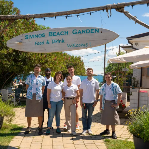 Group of people in casual uniforms standing under a sign at a garden cafe entrance.