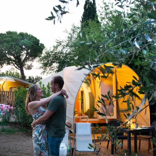 Couple embraces in a garden near a glowing tent at dusk.