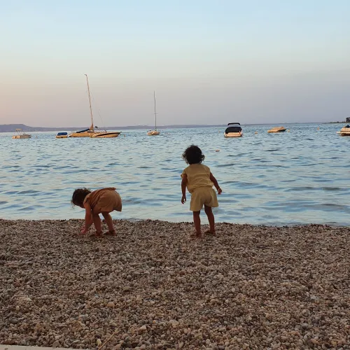 Two children play on a pebbled beach at sunset, boats in the background.