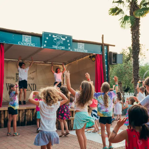 Children dancing in front of an outdoor stage with red curtains.