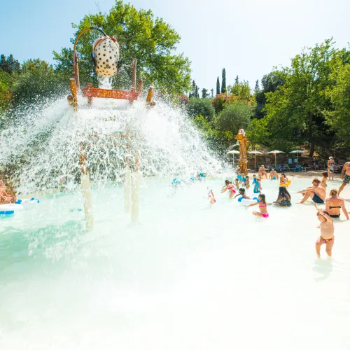 Children and adults play in a sunlit water park with falling water from a bucket.
