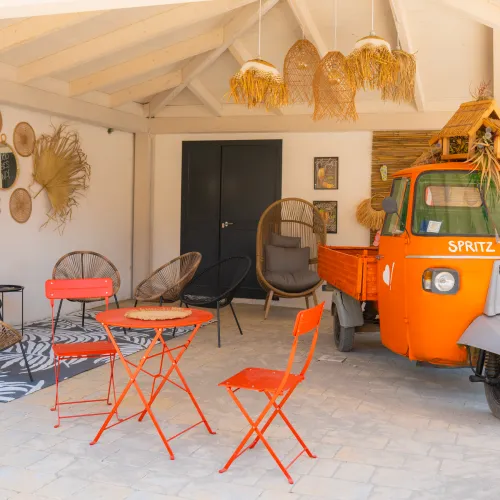 Orange vintage truck in a cozy patio with wicker decor and red metal chairs.